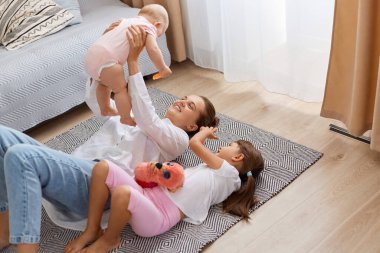 Portrait of happy mother and children playing in the living room resting on the floor, mommy raising arms and holding infant baby, elder sister lying near her family.