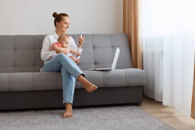 Portrait of happy woman in white t shirt sitting on sofa with baby daughter and working on laptop, holding smart phone in hands, searching information or chatting with friends.