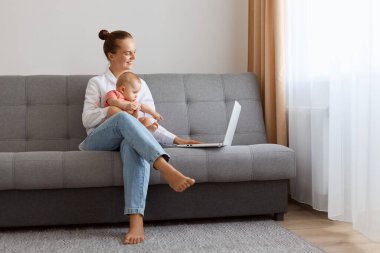 Indoor shot of smiling satisfied woman in white t shirt sitting on sofa with toddler daughter and working on laptop, looking at display of notebook with positive expression.