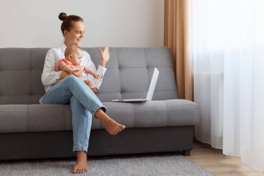 Indoor shot of smiling woman in white t shirt sitting on sofa with baby daughter and working on laptop, having video call, saying hi and showing hello gesture with hand.