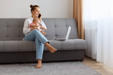 Attractive young woman with bun hairstyle in white t shirt sitting on sofa with baby daughter, female freelancer working on laptop and talking with partner on mobile phone.