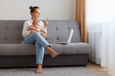 Serious concentrated beautiful woman in white t shirt sitting on sofa with baby daughter and working on laptop, young female works online during maternity leave.