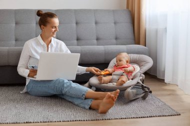 Image of delighted busy woman in white t shirt sitting near cough with baby daughter in rocking chair and working on laptop, typing on notebook and showing toy to her kid.
