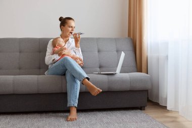 Angry Caucasian woman in white t shirt sitting on sofa with baby daughter and working on laptop, freelancer having troubles with online work, recording voice messages, screaming.