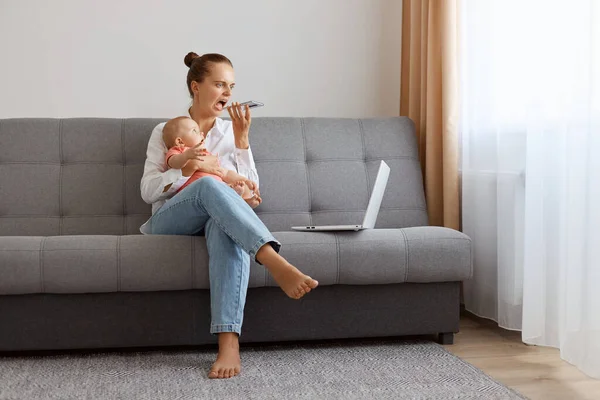 Angry Caucasian woman in white t shirt sitting on sofa with baby daughter and working on laptop, freelancer having troubles with online work, recording voice messages, screaming.