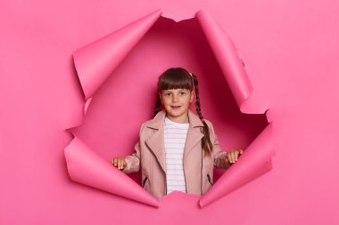 Indoor shot of positive optimistic little girl wearing jacket posing in pink paper torn, looking at camera with smile, expressing happiness, being in good mood.