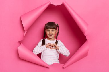 Indoor shot of charming excited little girl wearing striped shirt posing in pink paper torn, standing looking at camera, showing heart gesture with her palms.