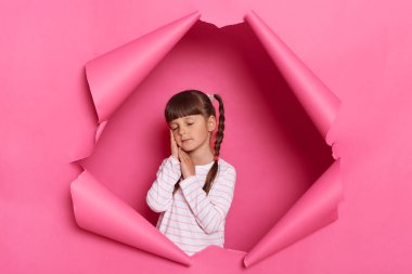 Indoor shot of sleepy tired little girl wearing striped shirt posing in pink paper torn, standing with closed eyes, having hap, leaning in her palms, being sleepless.