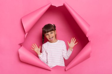 Portrait of happy positive funny little girl wearing striped shirt posing in pink paper torn, standing with raised arms and showing tongue out, having fun, frowning face.