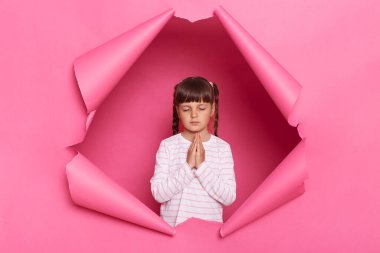 Indoor shot of calm little girl wearing striped shirt posing in pink paper torn, standing with closed eyes, keeping palms together in praying gesture.