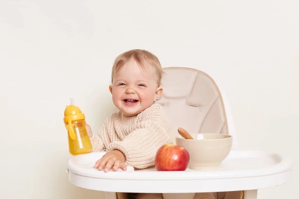 Indoor shot of smiling positive baby sitting in a chair against white isolated background, kid with water bottle and fruit having fun during breakfast or dinner.