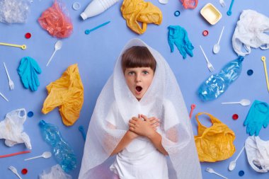 Horizontal shot of shocked stressed little girl posing against blue wall and much plastic garbage around, being astonished of planet pollution, looking at camera with big eyes.