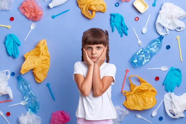 Indoor shot of depressed little girl wearing white t shirt posing against blue wall and much plastic garbage around, keeps palms on cheeks, asks stop destroying our planet.