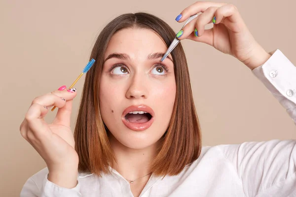 Indoor shot of concentrated woman with tweezers and eyebrow brush plucking brows hair, using metal forceps for eyebrows, posing with open mouth, looking up isolated over beige background.