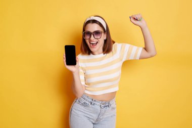 Portrait of happy woman wearing striped t shirt and jeans posing isolated over yellow background, showing blank black screen of smart phone and clenched fist, copy space for advertisement.