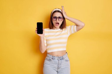 Shocked woman wearing striped t shirt and jeans posing isolated over yellow background, standing smart phone, showing empty display with copy space for promotional test, expressing astonishment.