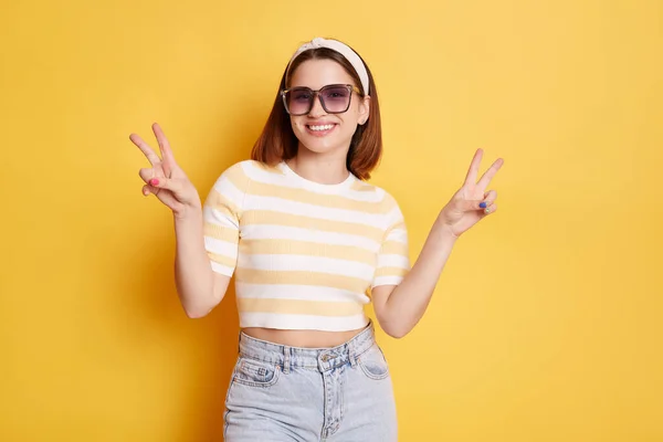 Horizontal shot of smiling delighted woman in striped t shirt and jeans, expressing positive emotions, showing v sign with both hands, celebrating her success, posing isolated over yellow background.