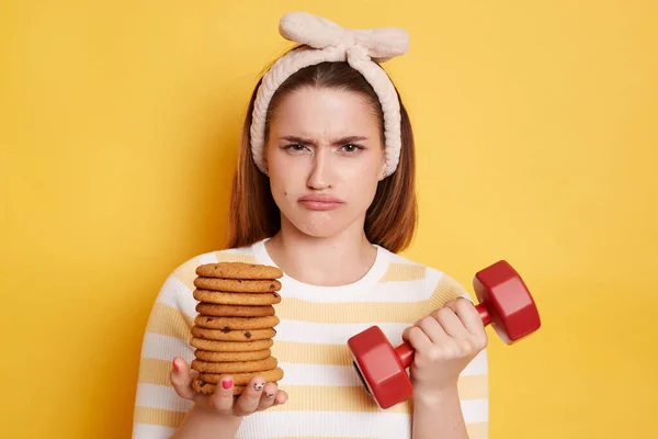 Portrait of upset sad woman with pout lips wearing striped shirt and hair band holding red dumbbell and cookies, needs to choose junk food or sport, posing isolated over yellow background.