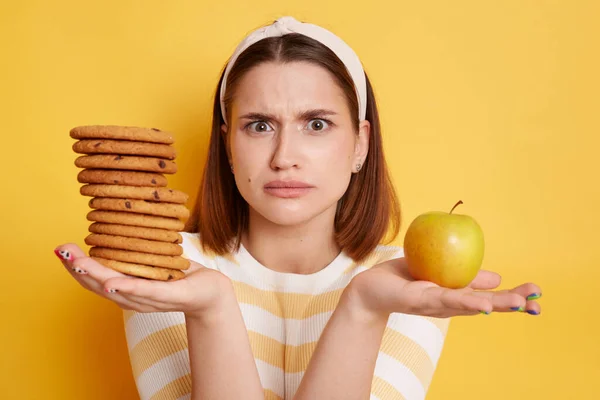 Portrait of confused woman wearing striped shirt and hair band, looking at camera with puzzlement, holding cookies and apple, does not know what to choose, posing isolated over yellow background.