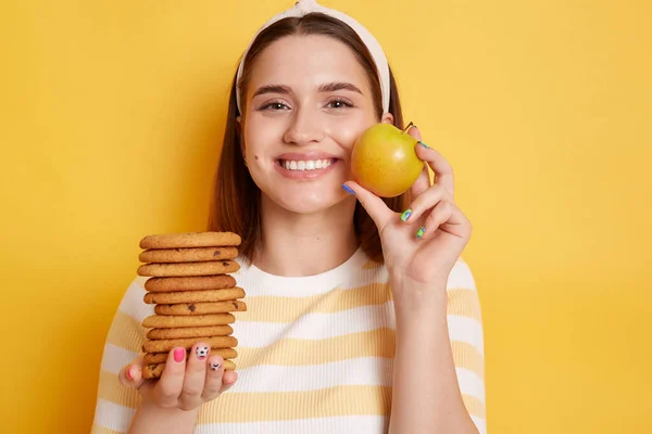 Attractive slip happy smiling woman wearing striped shirt and hair band, holding cookies and apple, prefer healthy eating instead of junk food, posing isolated over yellow background.