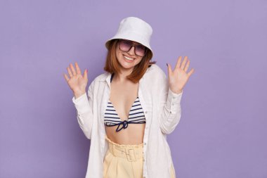 Portrait of good looking pretty young woman wearing panama, white shirt and swimming suit, posing isolated over purple background, standing with raised arms and toothy smile.