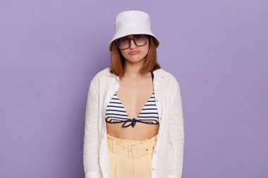 Image of young dark haired woman wearing panama, white shirt and swimming suit, being in bad mood, hearing unpleasant news, posing isolated over purple background.