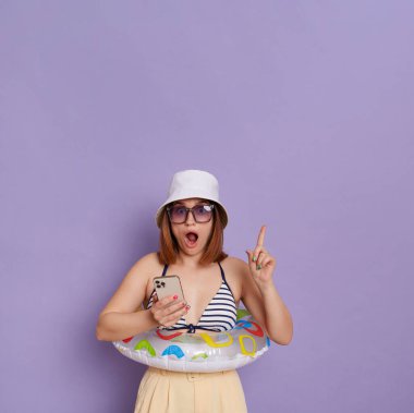 Image of shocked amazed woman with rubber ring wearing panama and swimming suit posing isolated over purple background, holding mobile phone and pointing finger at copy space, having idea.