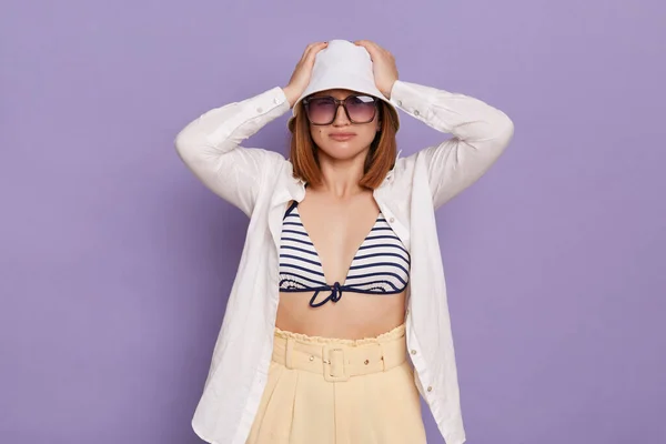 Image of sad upset woman wearing panama, white shirt and swimming suit, having problems with her memory, has troubles, keeps hands on head, posing isolated over purple background.