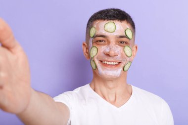 Portrait of smiling man wearing white t shirt applying mask with cucumber slices on his face, taking selfie, isolated over purple background, point of view photo.