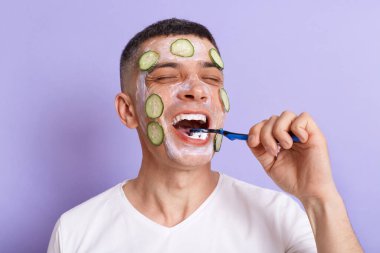 Positive happy man wearing white t shirt applying mask with cucumber slices on his face brushing his teeth, holding toothbrush in hands, keeps eyes closed, isolated over purple background.