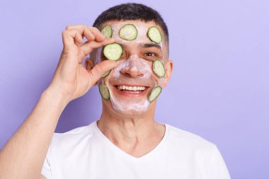 Image of positive joyful man wearing white t shirt applying mask, posing isolated over purple background, covering hid eye with cucumber slice, looking at camera with toothy smile.