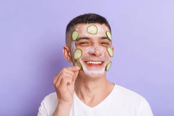 Horizontal shot of smiling satisfied man wearing white t shirt applying mask with cucumber slices on his face, enjoying cosmetology skin care procedures, isolated over purple background.