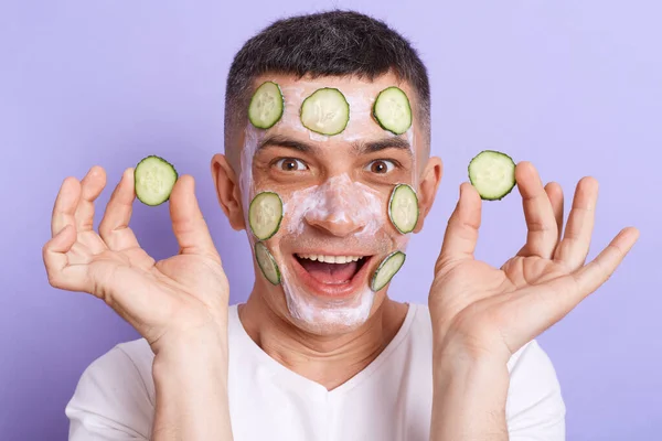 Image of shocked amazed young man wearing white t shirt applying mask with cucumber slices on his face and holding vegetable in hands, doing skin care procedures, isolated over purple background.