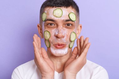 Indoor shot of pleasantly surprised man wearing white t shirt applaying mask with cucumber slices on his face isolated over purple background, being amazed of result of skin care procedures.