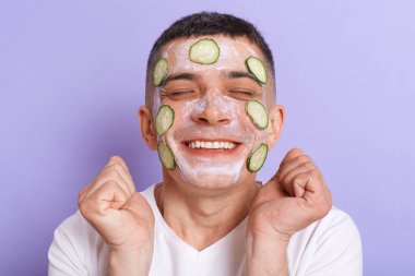 Extremely happy satisfied man wearing white t shirt applying mask with cucumber slices on his face, standing with clenched fists and closed eyes, posing isolated over purple background.