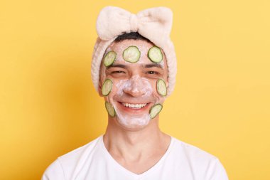 Happy young man wearing white t shirt and hair band doing cosmetic procedures with cream and cucumber slices on face, looking at camera with toothy smile isolated over yellow background.