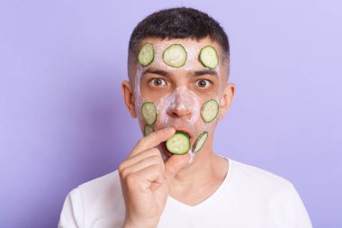 Horizontal shot of shocked man looking at camera with big eyes wearing white t shirt applying mask, covering mouth with cucumber slices isolated over purple background.