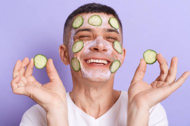 Portrait of extremely happy smiling man wearing white t shirt applying mask, holding cucumber slices in hand and put them on his face isolated over purple background.