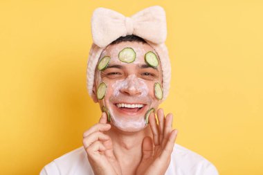 Smiling cute man wearing white t shirt and hair band doing cosmetic procedures with cream and cucumber slices on face, keeps hands on cheek, looking at camera, isolated over yellow background.