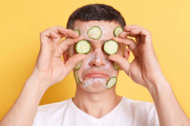 Sad upset man wearing white t shirt doing cosmetic procedures with cream, covering eyes with cucumber slices, standing with pout lips, expressing sadness, isolated over yellow background.