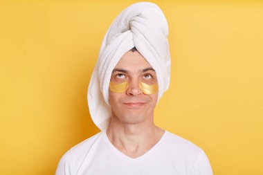 Portrait of pensive confused man wearing white t shirt and towel, having cosmetic patches under his eyes, looking up with puzzled facial expression, posing isolated over yellow background.
