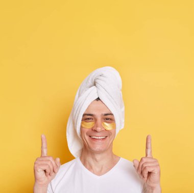 Satisfied man wearing white t shirt and towel, having cosmetic patches under his eyes, pointing up with fingers, showing space for advertisement, posing isolated over yellow background.