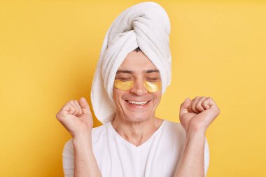 Indoor shot of positive satisfied man wearing white t shirt and towel, having cosmetic patches under his eyes, clenched fists, being extremely happy, posing isolated over yellow background.