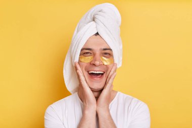 Excited happy man wearing white t shirt and towel, having cosmetic patches under his eyes, standing with palms on cheeks, expressing positive emotions, posing isolated over yellow background.