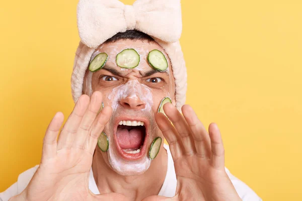 Angry man wearing white t shirt and hair band doing cosmetic procedures with cream and cucumber slices, making announcement, screaming loud, isolated over yellow background.