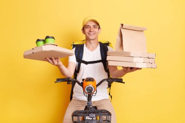 Indoor shot of positive optimistic courier wearing cap and white t shirt holding pizza boxes and coffee from coffee, riding bicycle, delivering food, isolated on yellow background