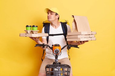 Horizontal shot of courier wearing cap and white t shirt holding pizza boxes and takeaway drink, riding bicycle, posing isolated on yellow background, smelling and enjoying tasty coffee.