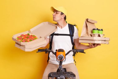 Deliveryman wearing cap and white t shirt holding pizza boxes and coffee, riding bicycle, smelling delicious order with closed eyes and enjoying, posing isolated on yellow background