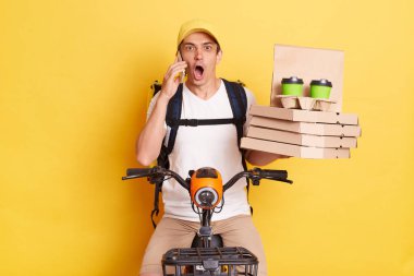 Horizontal shot of shocked astonished courier man holding mobile phone, talking with client or boss, being very surprised, wearing cap and thermo bag, riding bicycle, isolated on yellow background