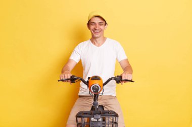 Indoor shot of smiling cheerful man wearing white t shirt and cap riding electric scooter, having fun alone while having free time, isolated on yellow background.
